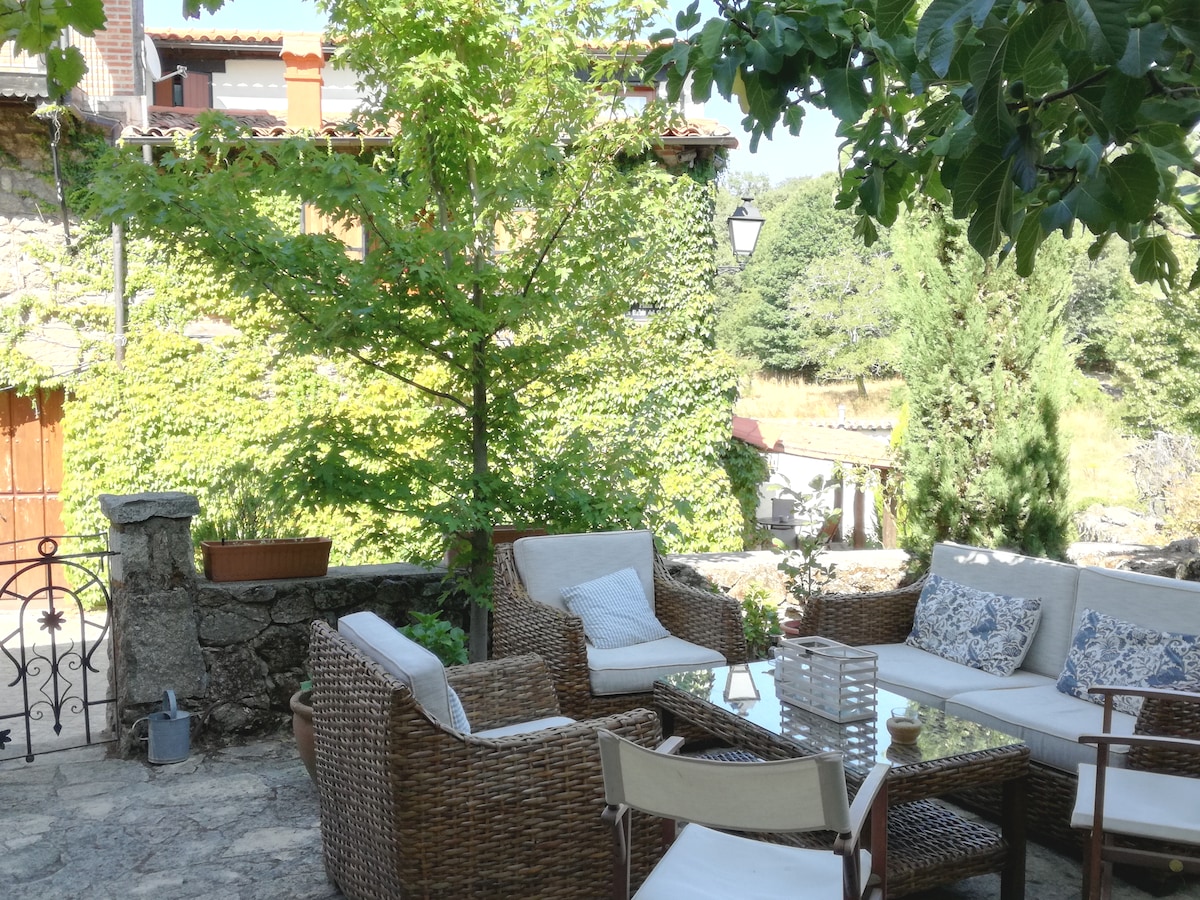 A comfortable outdoor seating area showcases woven furniture arranged around a glass-top table, set against a backdrop of lush greenery. A young tree provides shade, while the stone wall adds rustic charm to the inviting space.