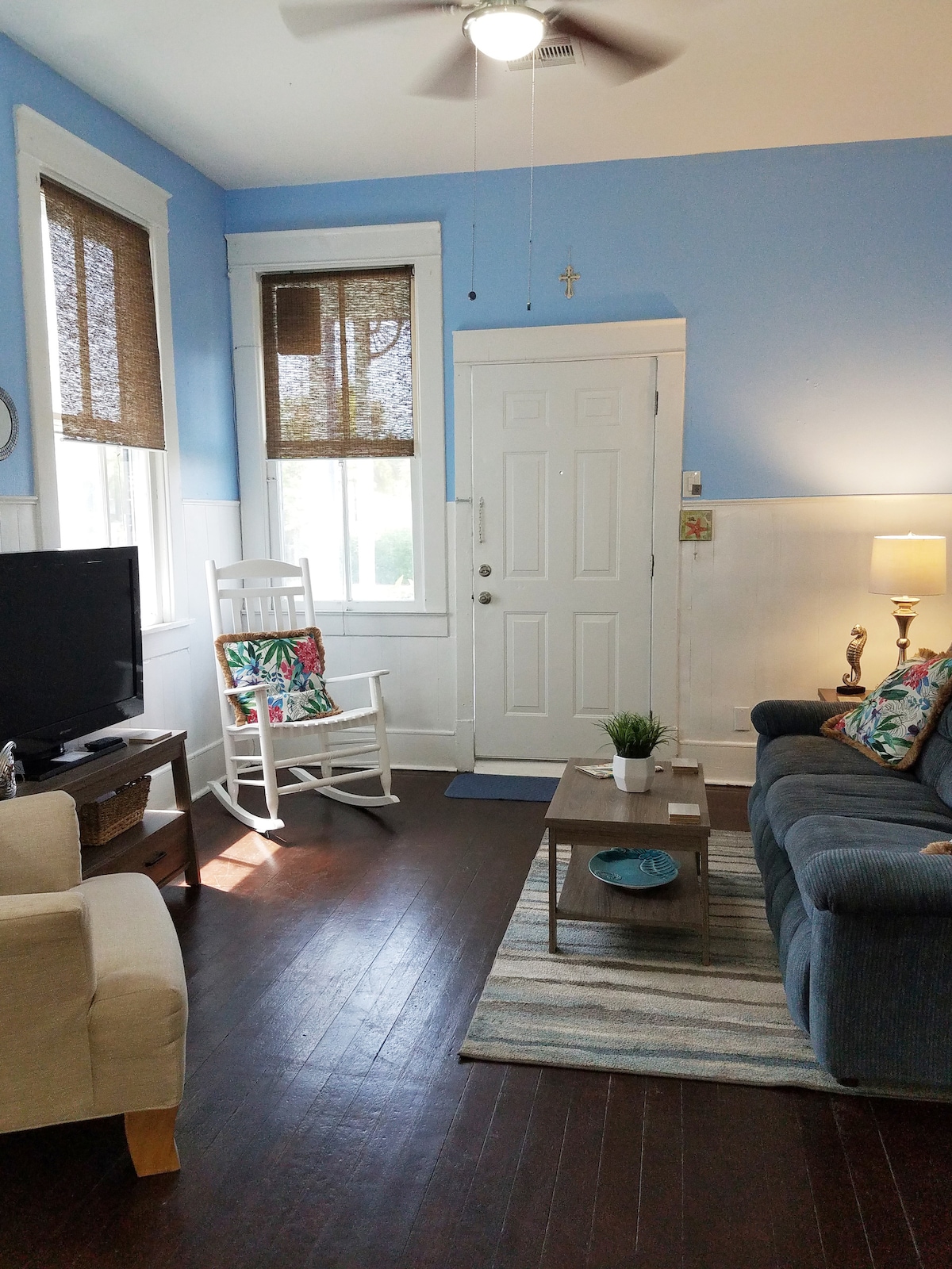 The living room features hardwood floors and high ceilings, illuminated by natural light from the windows. A comfortable sofa is paired with a cozy armchair, and a small coffee table is centered on a striped area rug. A television and lamp provide additional amenities.
