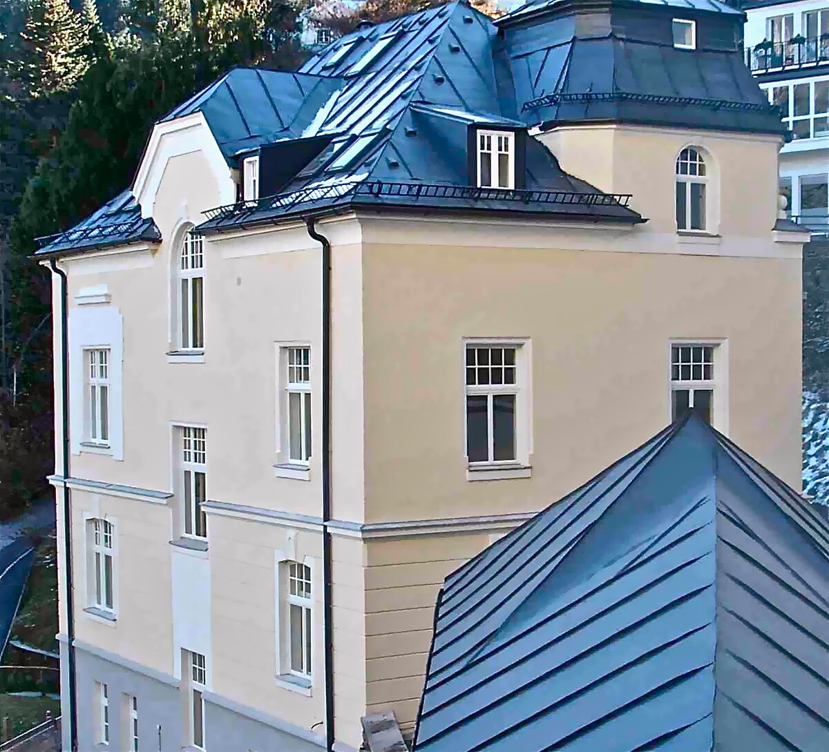 The exterior of a building is presented, highlighting its elegant architectural design. The structure features a blend of light-colored walls and dark metal roofing, with multiple windows that allow for ample natural light. Nearby trees and hillside are visible in the background.
