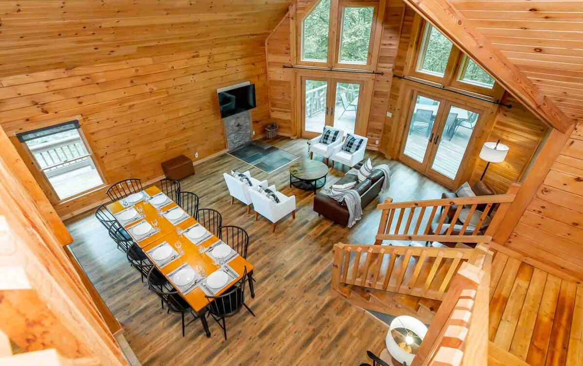 An airy great room showcases high wooden ceilings and large windows that allow natural light to fill the space. A long dining table is set for meals, and comfortable seating arrangements are positioned around a coffee table. A staircase leads to the upper level.