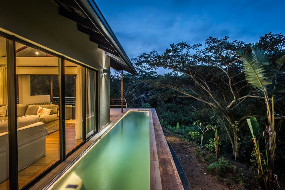 The image captures a sleek swimming pool extending from a modern home, bordered by a wooden deck. Large glass doors provide a view of a spacious lounge area, with soft furnishings illuminated by the evening light. Indigenous trees and greenery are visible in the background.