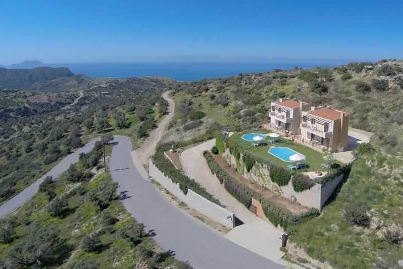 An aerial view captures the two adjacent villas, each with private pools, set against a backdrop of rolling hills and the sea. A winding road leads to the villas, which are surrounded by landscaped gardens and open spaces. The blue sea stretches in the distance.