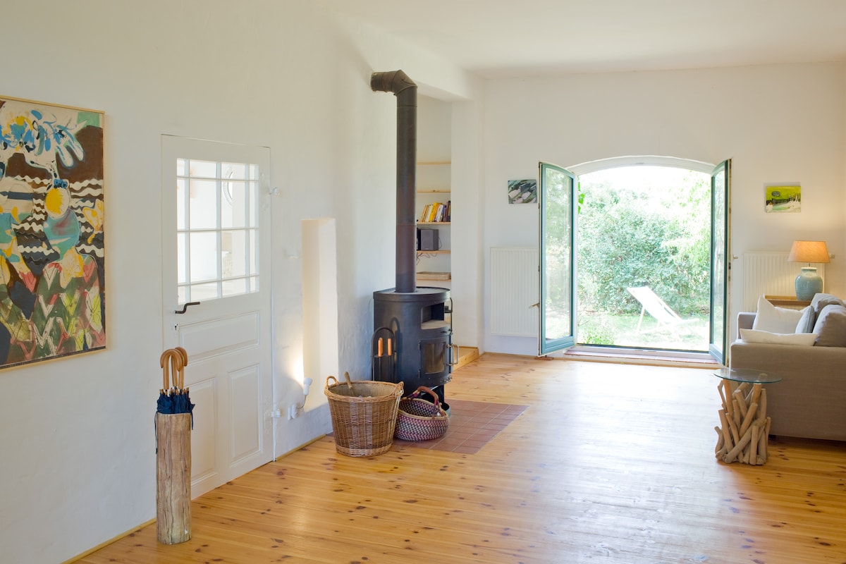 A spacious living area features a wood-burning stove, light wooden flooring, and white walls. Open doors reveal a glimpse of the garden outside. A comfortable sofa and a soft lamp provide cozy seating. Colorful wall art adds visual interest to the bright space.