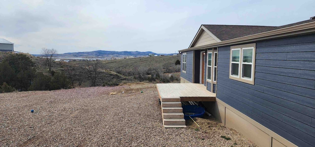 A side view of the house reveals wooden steps leading to a deck area, surrounded by a gravel landscape. The blue exterior contrasts with the natural surroundings, while distant hills and fields are visible in the background, enhancing the home's private setting.