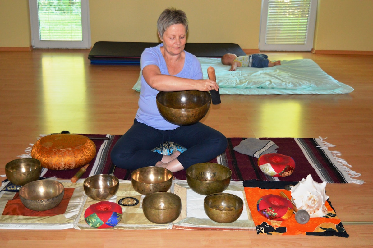 A wellness practitioner sits cross-legged on a wooden floor, focused on playing a singing bowl. Several bowls of varying sizes are arranged neatly around her, each placed on colorful mats. A soft mat can be seen in the background, along with large windows allowing natural light to fill the space.