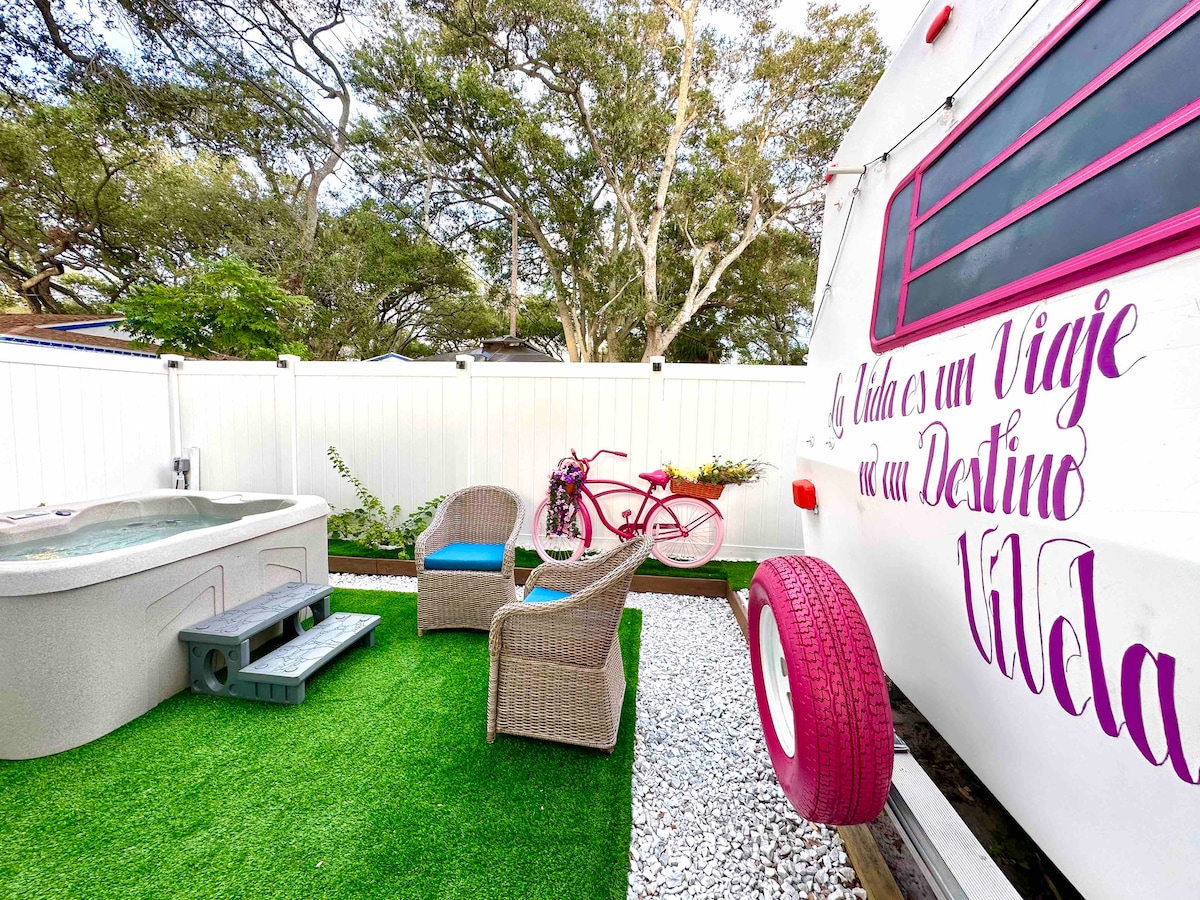 A private outdoor area features a hot tub and two wicker chairs placed on vibrant green grass. A decorative bicycle in bright pink adds a pop of color beside the white privacy fence. Gravel pathways enhance the serene atmosphere of the space.