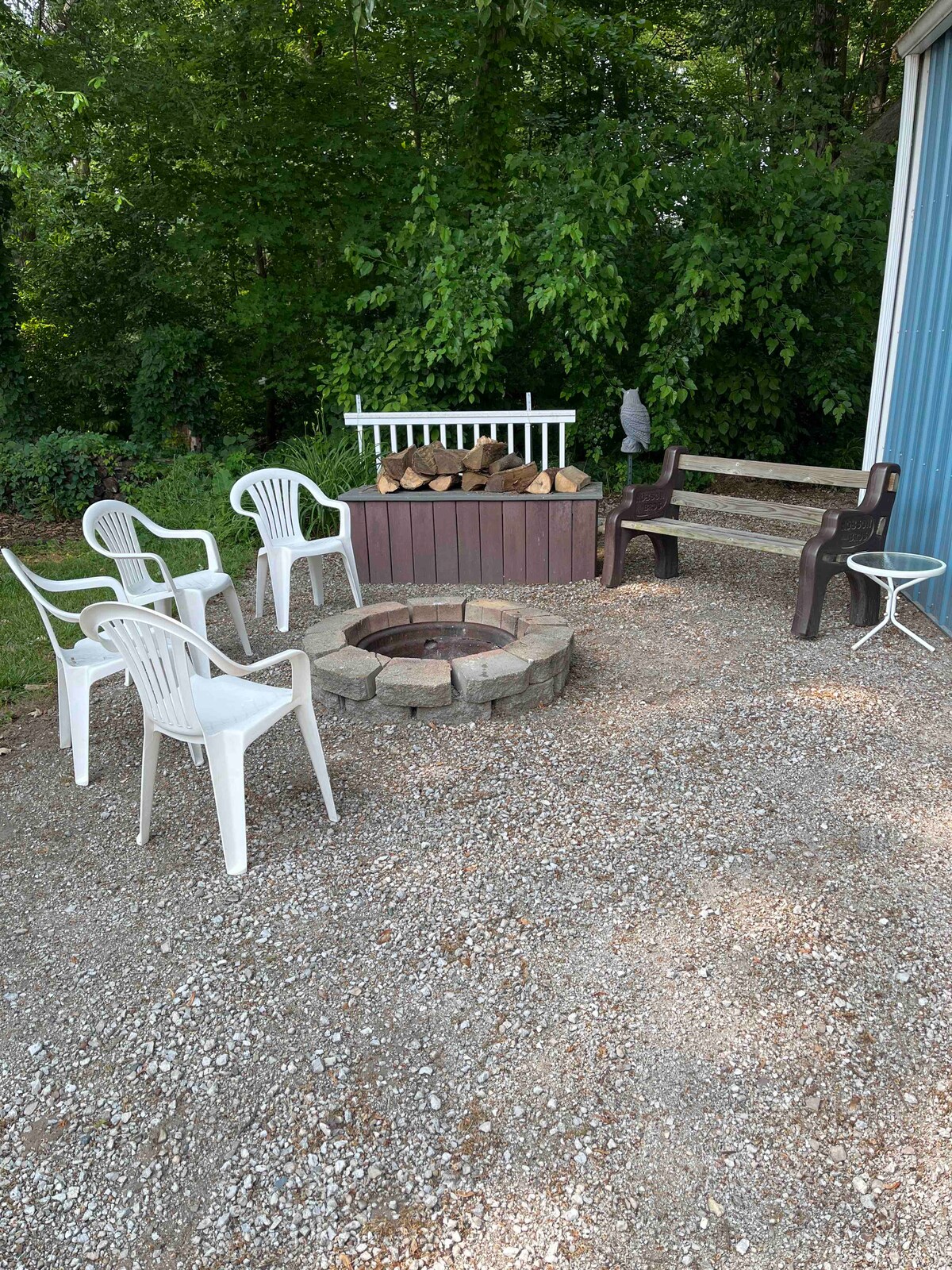 An outdoor gathering area is presented with a stone fire pit surrounded by four white chairs and a brown bench. Nearby, a stack of firewood is neatly arranged. Lush greenery offers privacy and a calming backdrop.