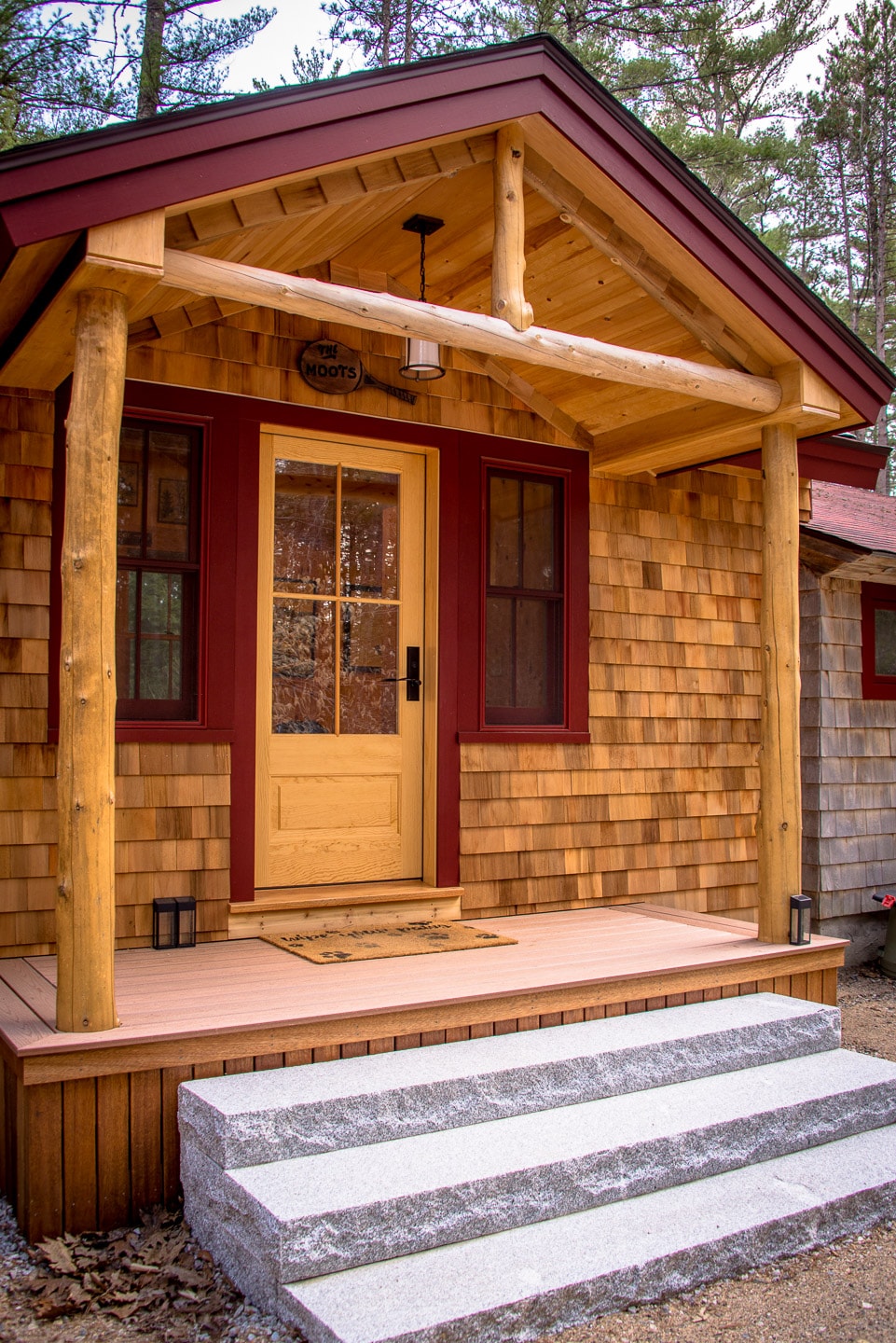 A welcoming cabin entrance features natural wood siding complemented by deep red accents. A sturdy yellow front door is framed by a wooden overhang, while stone steps lead up to a small porch, providing a charming focal point surrounded by trees.