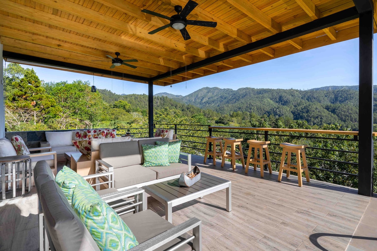 An outdoor terrace presents comfortable seating with neutral-colored sofas adorned with green accents. Sturdy wooden stools complement the area, while panoramic mountain views create a serene backdrop. The space is shaded by a wooden ceiling, enhancing the connection to nature.