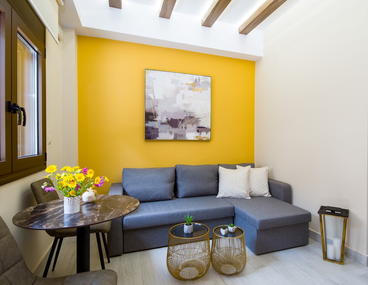 A cozy seating area is presented with a grey sectional sofa and a round marble table. A vibrant yellow wall adds brightness to the space. A floral arrangement and small decorative items are positioned on the table, enhancing the room's welcoming appeal.