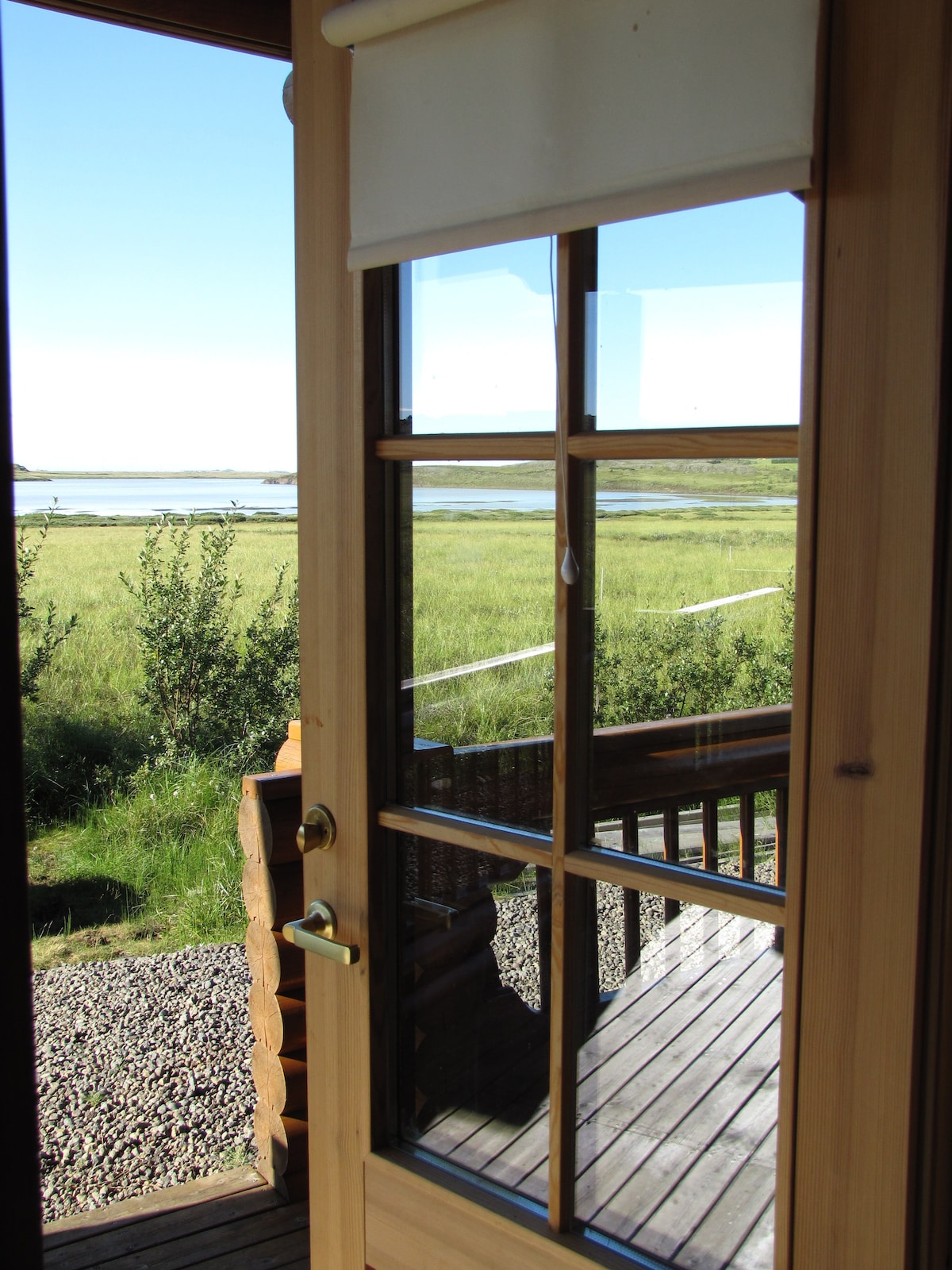 A wooden door with glass panels opens to reveal a view of a lush green landscape and a river. Sunlight illuminates the scene, and decking is visible underfoot. The natural surroundings create a peaceful atmosphere.