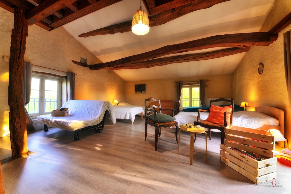 A spacious room features exposed wooden beams across the ceiling, complemented by warm lighting. Multiple beds are arranged thoughtfully, along with a chair and small table. Large windows permit natural light, illuminating the neutral-toned walls and wooden floor, creating an inviting and open atmosphere.