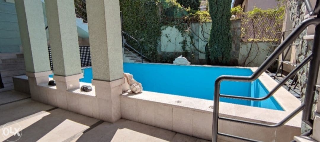 A rectangular swimming pool with clear blue water is framed by smooth stone edges. Steps are present on one side for easy access, while lush green foliage is observed in the background, providing a sense of privacy and natural beauty.