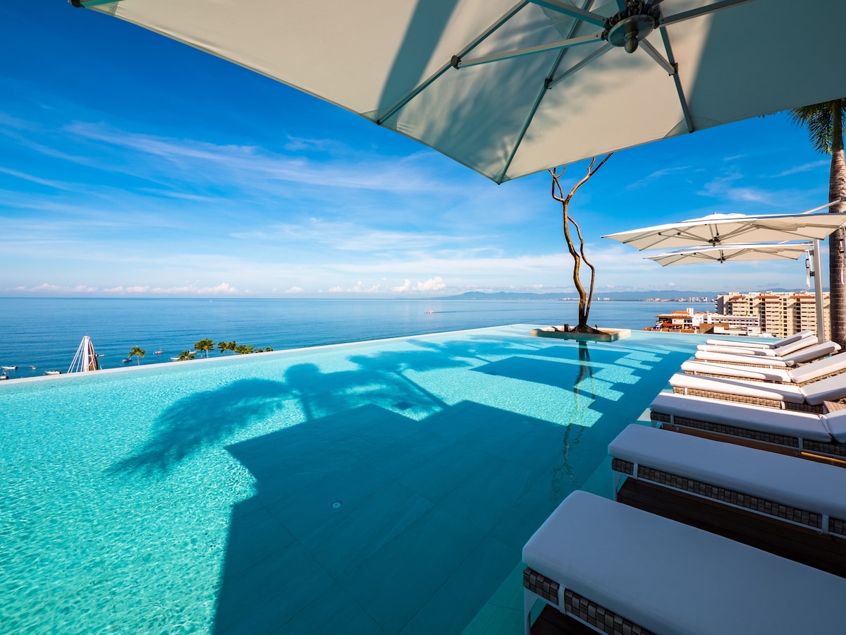 An infinity pool extends horizonally, blending into the ocean views. Sun loungers are positioned along the pool's edge, shaded by large umbrellas. A single tree stands near the pool, casting shadows on the water as the sky displays clear blue tones.