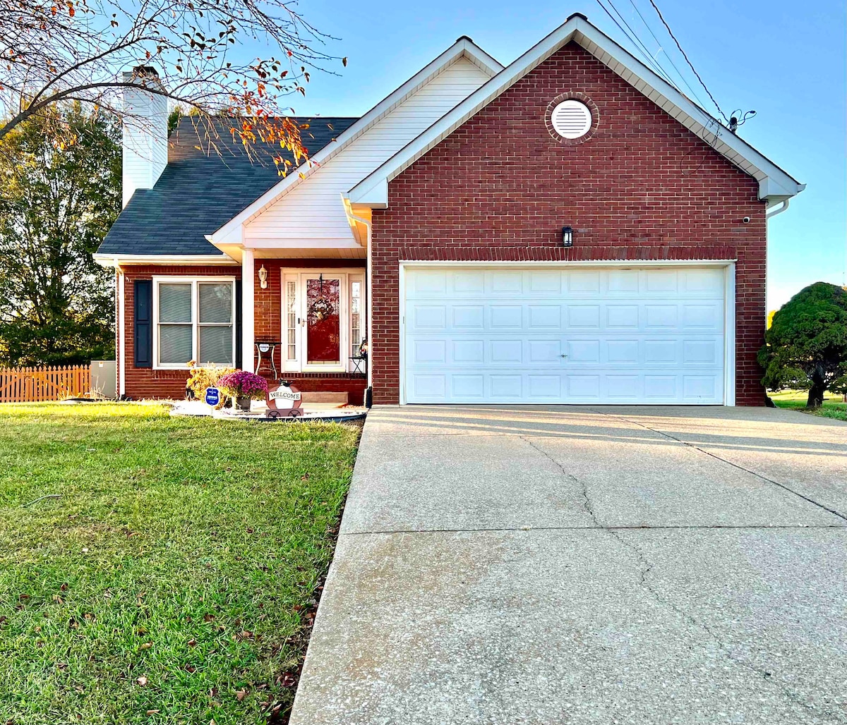 The front exterior of the home features a brick facade complemented by a white garage door. A well-maintained lawn is visible, with colorful flowers and shrubs arranged near the entrance. Trees provide greenery to the surroundings, creating a welcoming appearance.