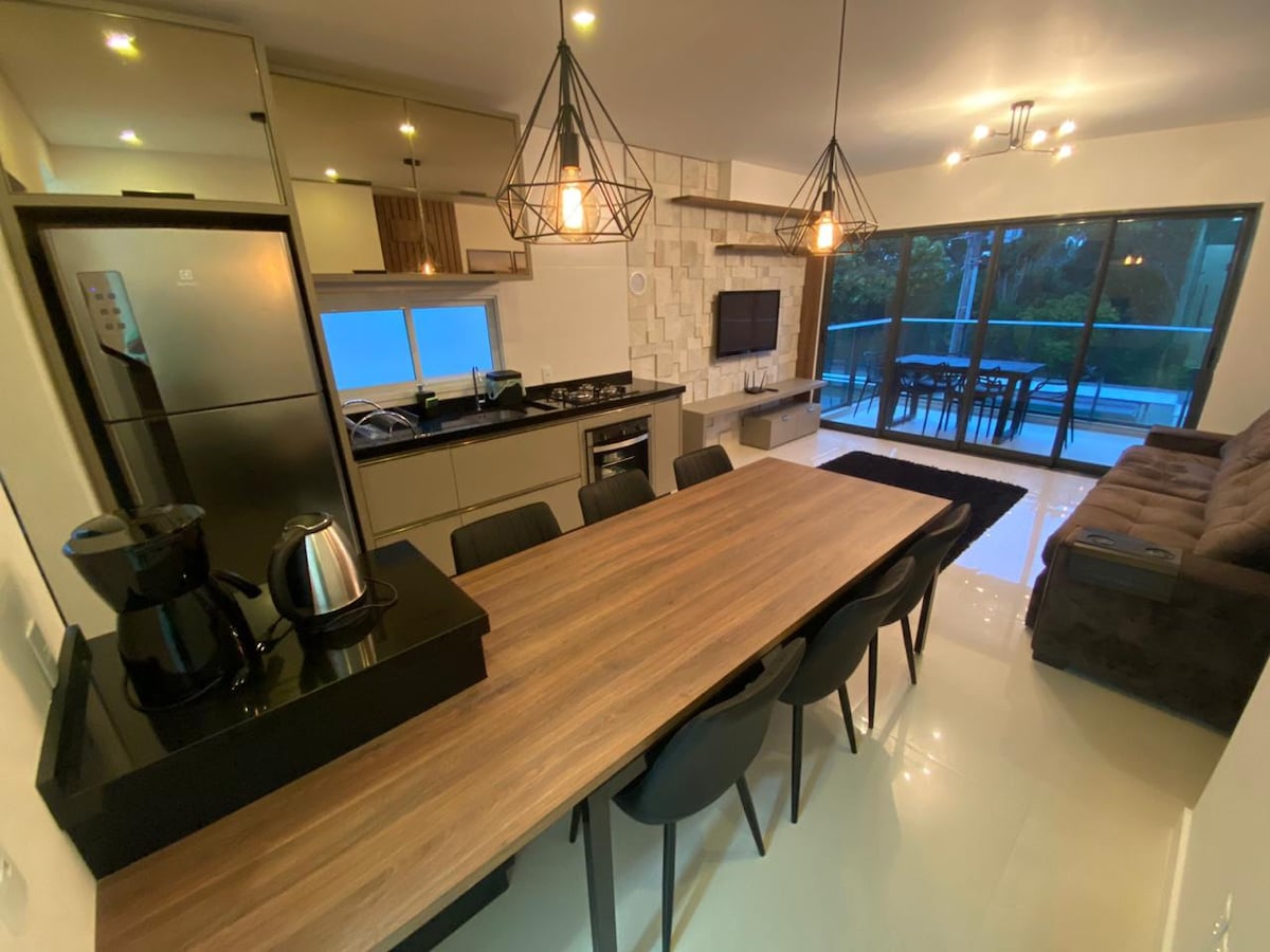 A modern open-concept kitchen and dining area features a long wooden table surrounded by black chairs. Sleek appliances line the kitchen, and large glass sliding doors provide views of the outdoor space and allow natural light to fill the interior.