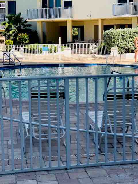 A swimming pool area is visible, featuring clear blue water surrounded by a paved deck. Several lounge chairs are positioned near the pool, offering a space for relaxation. A secure fence encloses the pool for safety, with lush greenery in the background.