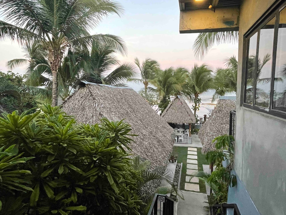 A view of a tropical landscape showcases thatched-roof structures surrounded by greenery. Palm trees are present, creating a tranquil environment, while the beach is visible in the background, hinting at a nearby river mouth and inviting outdoor seating areas.
