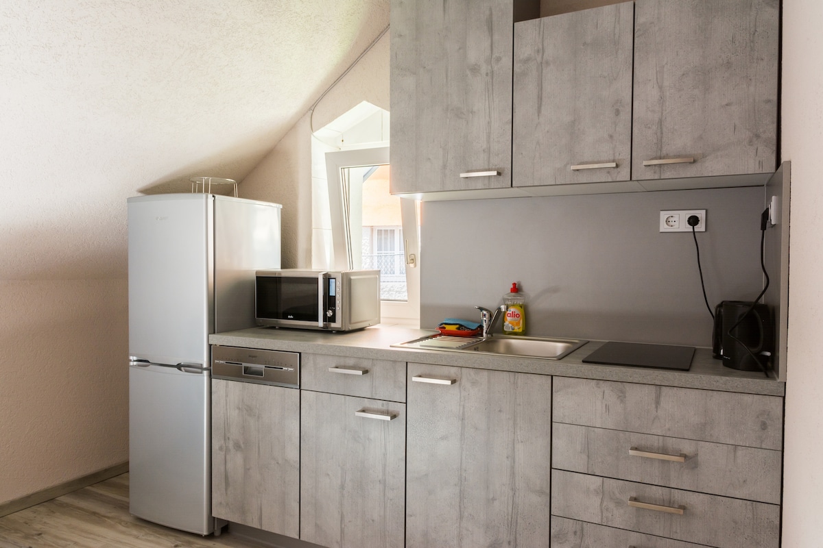 A functional kitchen area is presented with modern cabinetry in a light grey finish. It includes a refrigerator, microwave, and stovetop. A window allows natural light to enter, while various kitchen utensils are neatly arranged on the countertop.