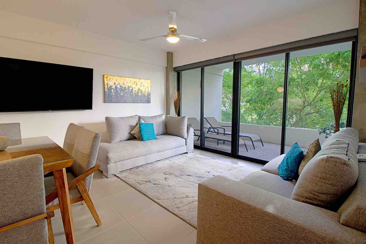 A modern living space features a comfortable seating area with light gray sofas, complemented by a textured area rug. A wooden dining table with chairs is positioned nearby. Floor-to-ceiling windows allow natural light and provide views of the greenery outside.