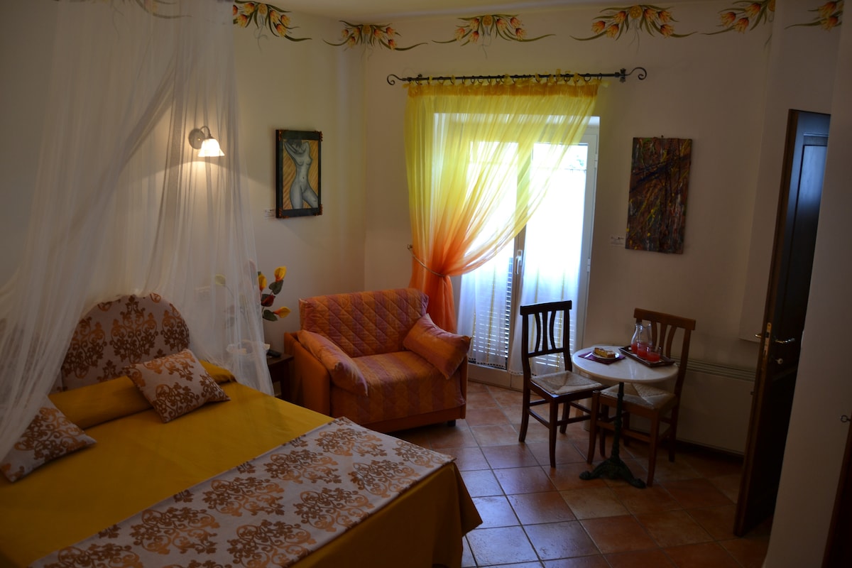 A warmly decorated bedroom features a spacious bed with an orange and yellow color scheme. Light flows through a window dressed in sheer fabric. A cozy seating area is placed nearby, furnished with a patterned couch, two chairs, and a small table.