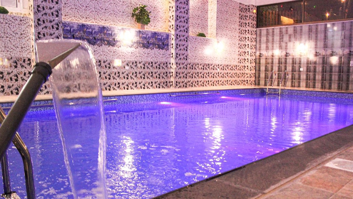 An indoor pool is seen, featuring a tranquil blue surface reflective of the soft lighting above. The pool area is highlighted by decorative wall panels and gentle water features, creating a serene environment for relaxation and leisure.