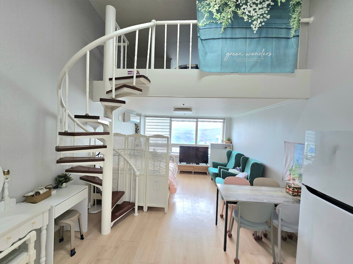 A spacious, open layout features a spiral staircase leading to an upper level with beds. A comfortable seating area includes teal chairs and a small dining table. Soft natural light fills the space, complemented by a simple decor with green accents.