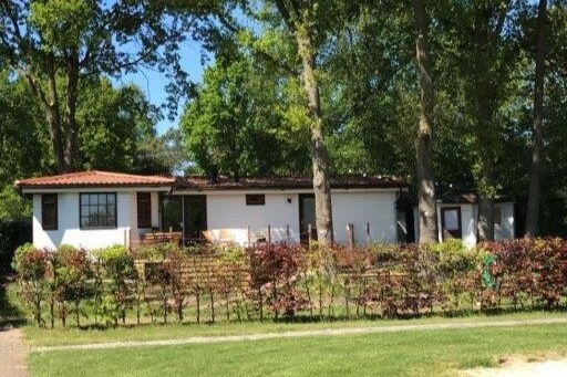 A spacious chalet is surrounded by greenery, featuring a wooden porch at the front. Large windows allow natural light to fill the interior, while a well-maintained garden with blooming plants provides a welcoming entrance. Trees offer shade and enhance the natural setting.