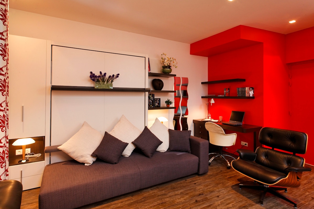 A contemporary studio is presented with a cozy seating area featuring a spacious dark couch adorned with multiple pillows. A sleek workspace is positioned against a vibrant red wall, with additional decorative elements adding character to the room. Warm lighting highlights the modern décor.