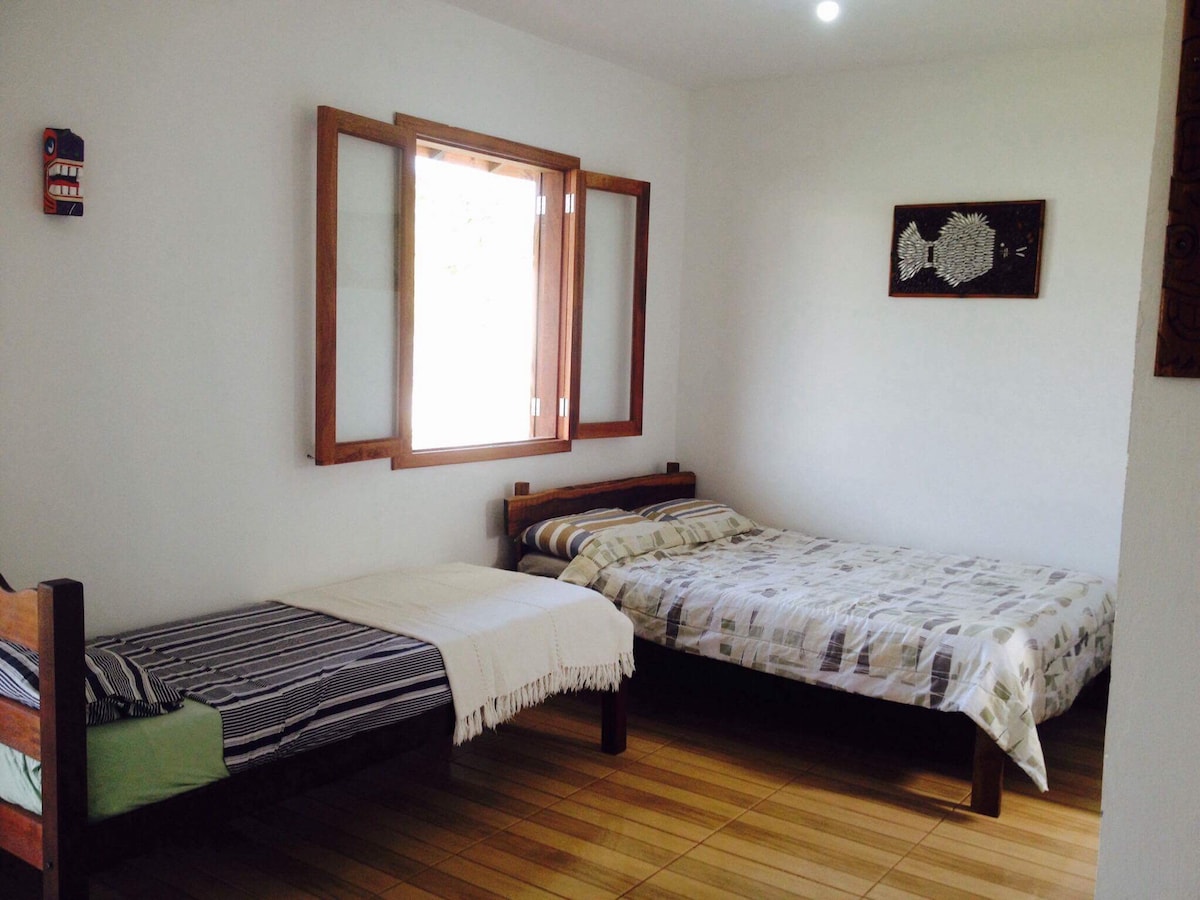 The sleeping area features two beds, one larger and one single, arranged on a wooden floor. A window with wooden frames allows natural light in, illuminating the light-colored bedding. A piece of wall art is displayed above the beds, adding a touch of decor.