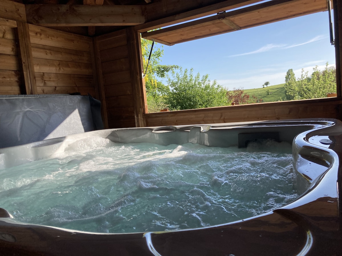 A spacious hot tub is set within a wooden structure, surrounded by natural wood paneling. Sunlight streams through an open window, illuminating the bubbling water while views of greenery are visible in the background.