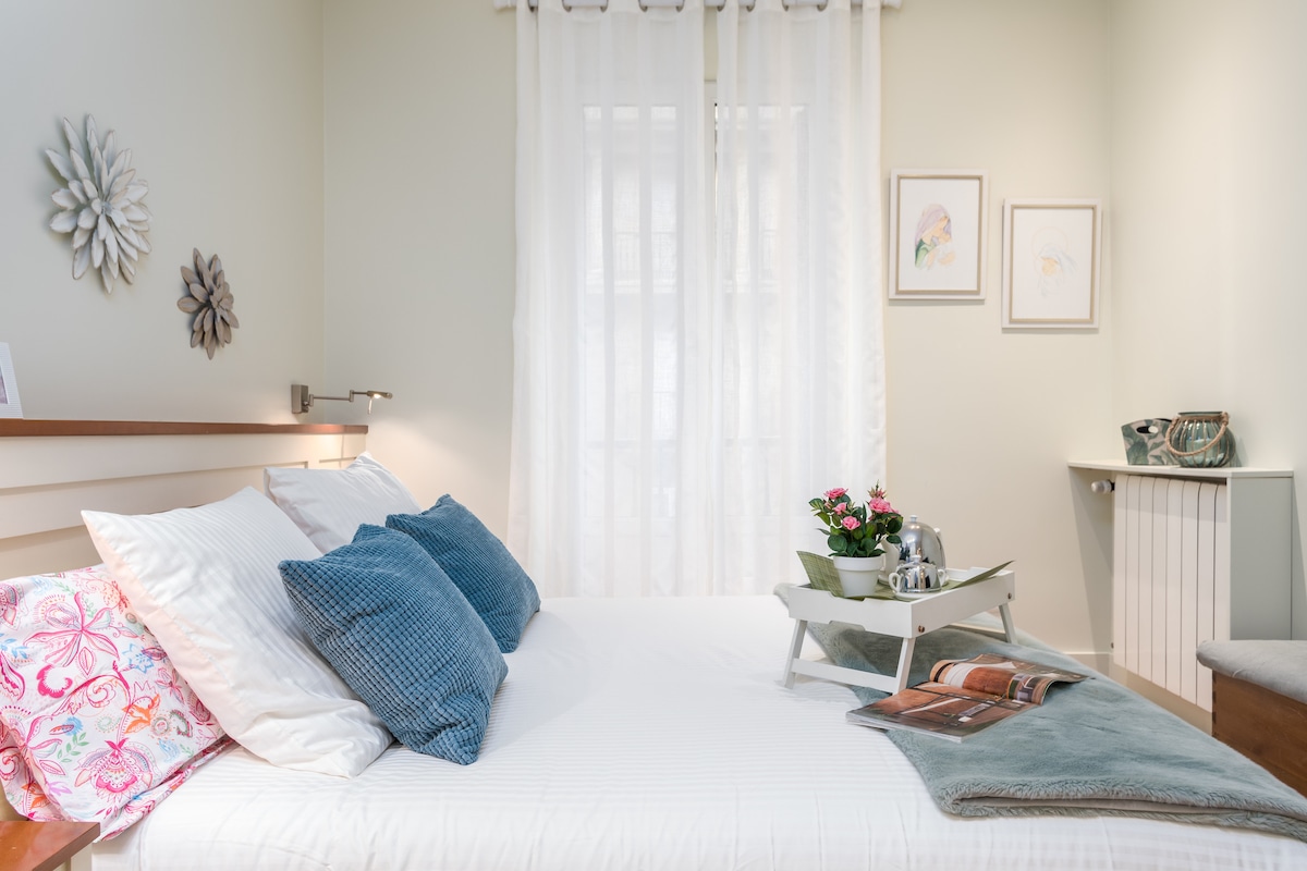 A bright bedroom features a spacious bed adorned with white linens and vibrant pillows. Natural light enters through sheer curtains. A bedside table holds a decorative tray with a teapot and flowers. Art pieces are displayed on the wall, contributing to a serene ambiance.