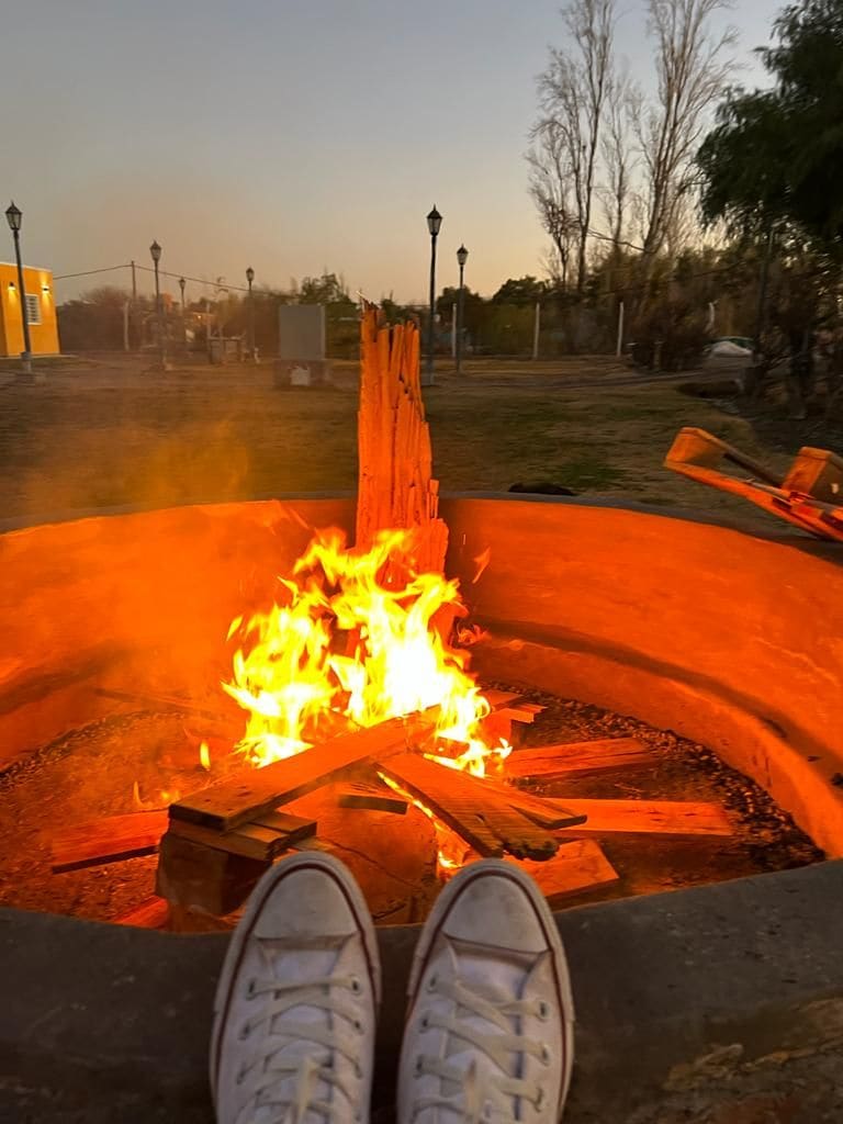 A fire pit is shown, with flames dancing above wooden logs. Warm orange and yellow hues illuminate the surroundings. The ground is adorned with gravel, and the silhouette of trees and other structures can be seen in the background, reflecting a tranquil outdoor environment.