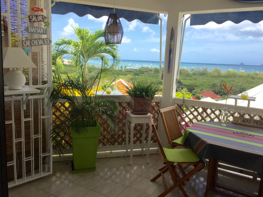 A covered terrace offers panoramic views of the Sainte Anne Bay, complemented by lush greenery. A small dining table and folding chairs are present, with a decorative plant adding a pop of color. The space is well-lit by natural light and features a ceiling lantern.