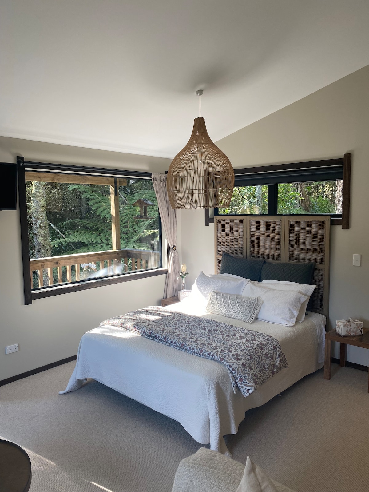 A serene sleeping area features a spacious bed dressed in soft linens. Large windows invite natural light and showcase views of the surrounding native bush. A decorative hanging light adds visual interest above the bed, complemented by a cozy throw and accent pillows.