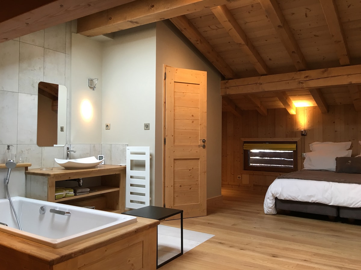 The spacious room features wooden beams and a warm color palette. A comfortable bed is positioned alongside a modern sink and wooden cabinetry. A bath with a sleek design is visible, alongside a large mirror. Natural light filters in through the window.