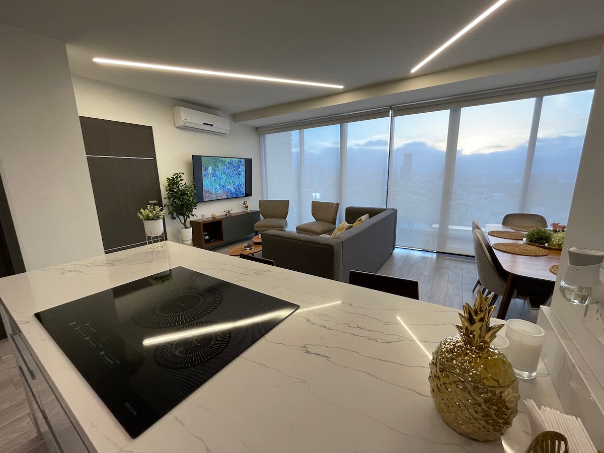A modern living area is visible with a comfortable seating arrangement and a large wall-mounted TV. The kitchen counter features an induction cooktop, while large glass windows provide a view of the surrounding area. Soft natural light filters through sheer curtains.