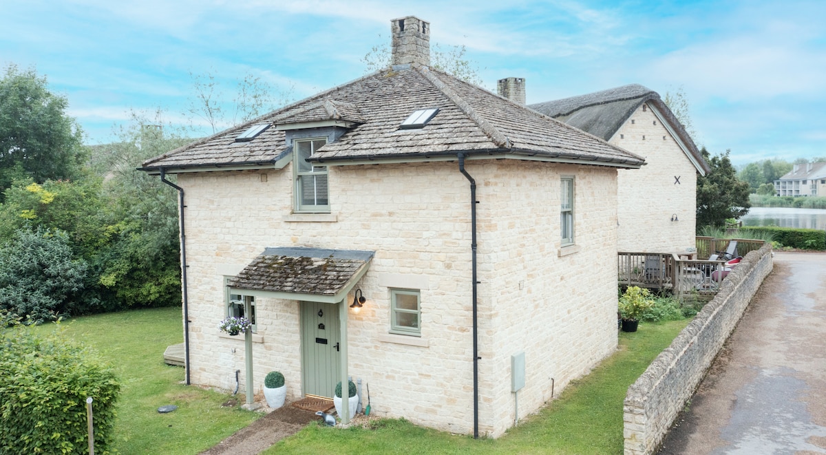 The cottage features local stone construction, showcasing a welcoming façade with a front door and a small porch. Greenery surrounds the property, offering a spacious lawn, while large windows let in natural light. The nearby structure and serene water backdrop enhance the setting.
