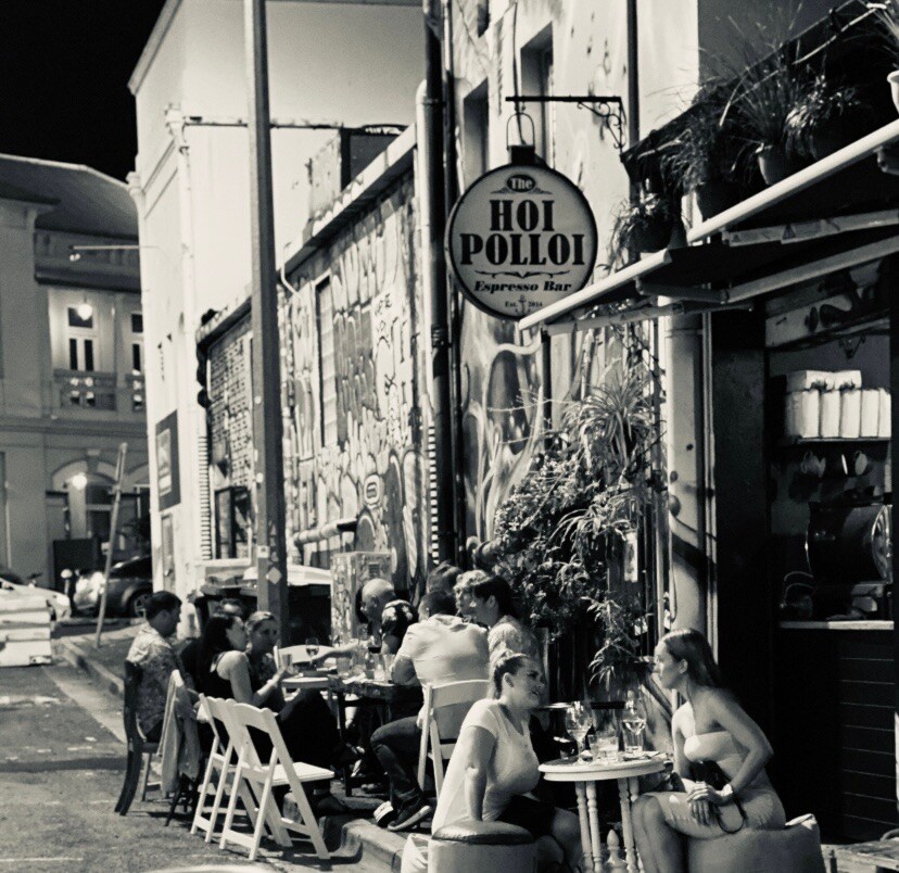 A lively street scene showcases outdoor dining with tables set for guests along a vibrant mural. Patrons are seated, enjoying their meals under ambient street lighting. An inviting cafe with a prominent sign adds to the bustling atmosphere, typical of an active urban environment.