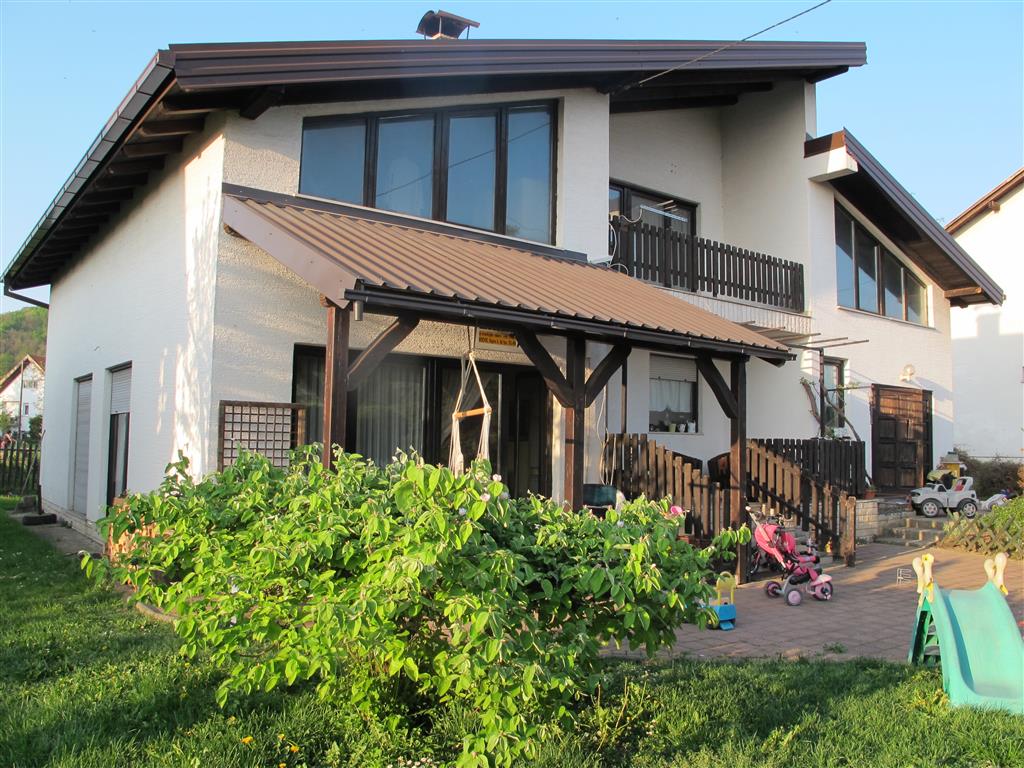 A two-story family house is depicted, featuring a covered entrance with wooden beams. The garden includes green grass and a flowering bush. Playground equipment, including a slide and toys, is visible in the yard, enhancing the outdoor space for family activities.