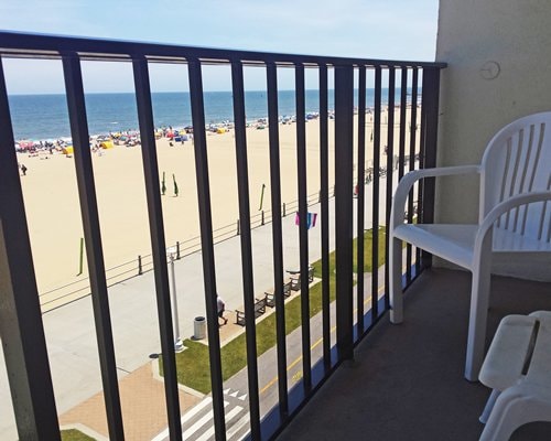 A private balcony is visible, featuring a white chair. The balcony overlooks a sandy beach and the ocean, where beachgoers are seen in the distance. A promenade with benches runs alongside the beach, providing a spacious view of the coastal scenery.
