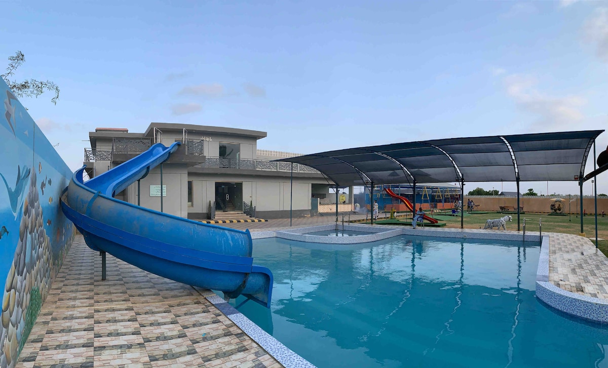 A vibrant swimming pool features a blue water slide, complemented by shaded seating areas. The pool is surrounded by tiled flooring, and a mural depicting marine life adds a playful touch. Lush green lawn borders the area, ensuring a refreshing and relaxing setting.