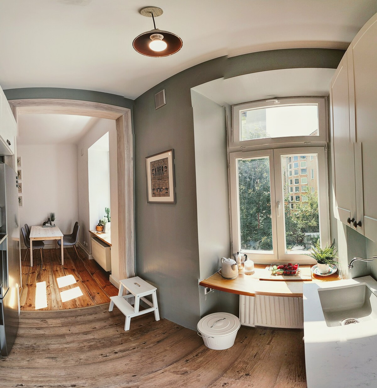 A bright kitchen area features light-coloured cabinets and a white countertop. Natural light streams in through a large window, illuminating a small dining table set against a wall. Wooden floors enhance the warm ambiance, while a stool and various potted plants are also visible.