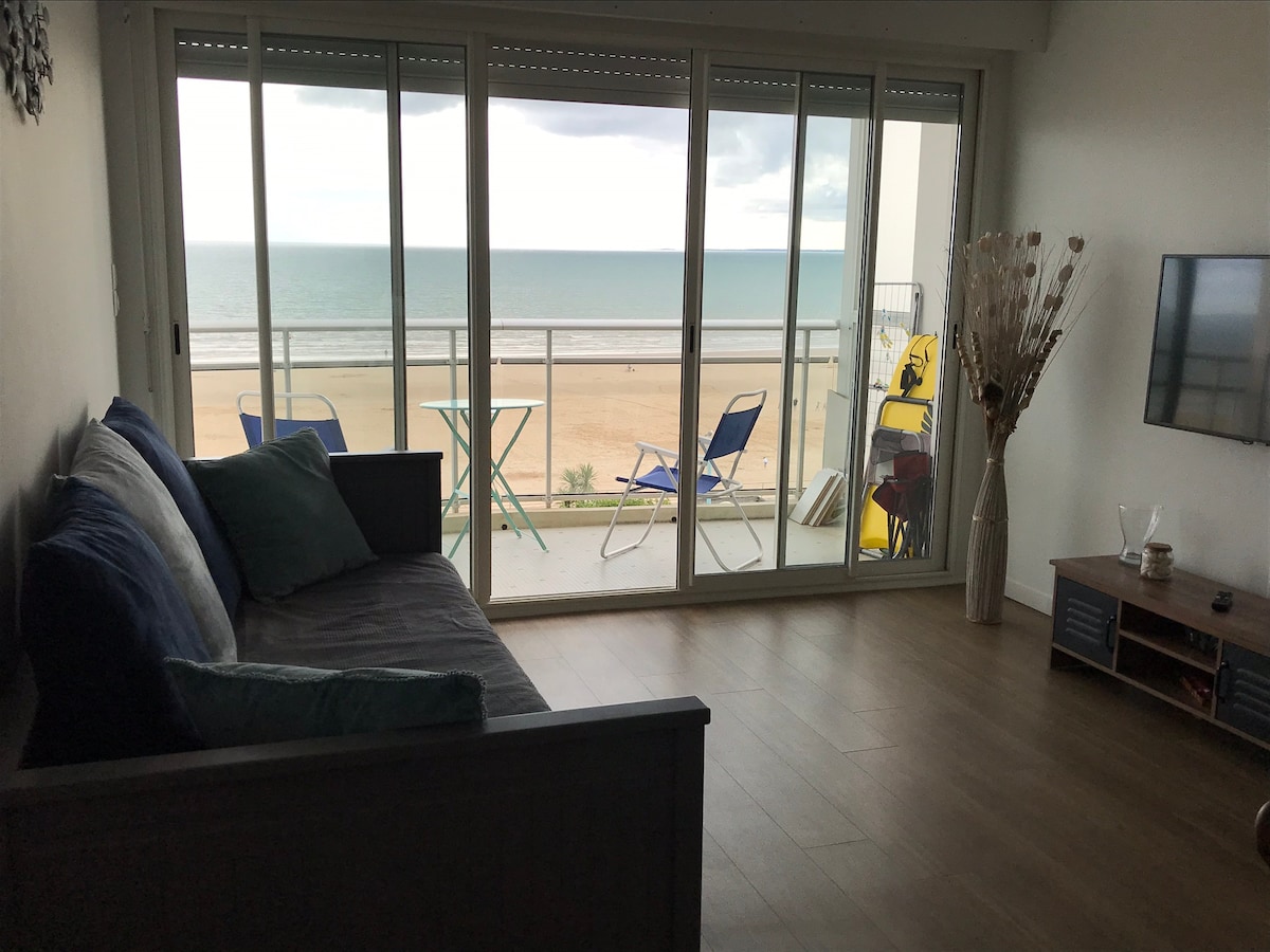 A spacious living area features large glass doors that open to a balcony overlooking the beach. Coastal views are visible beyond the outdoor seating. Soft furnishings in neutral tones are paired with a wall-mounted television, enhancing the inviting atmosphere.