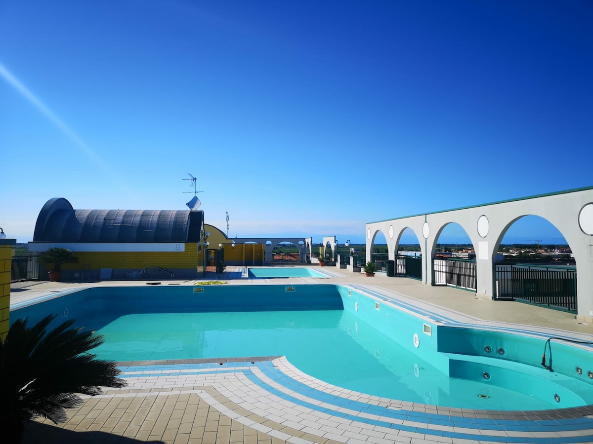 A panoramic pool area is shown, featuring clear turquoise water and surrounding sun terrace areas. Architectural arches provide an elegant backdrop, while a blue sky and distant sea view enhance the space's tranquil ambiance. A few palm plants can be seen near the edge of the pool.