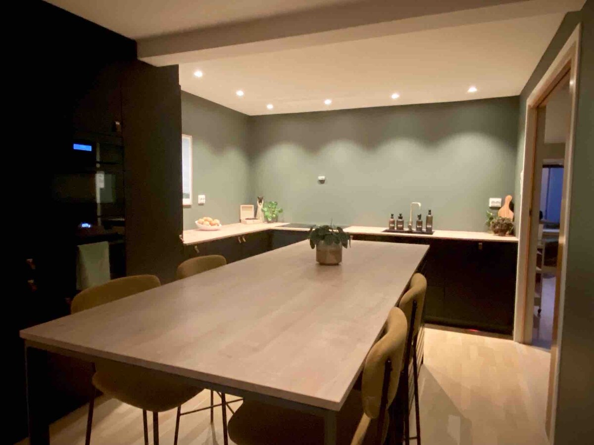 A modern kitchen features a long wooden table surrounded by several chairs. The space is illuminated with recessed lighting, and dark cabinetry complements the muted green walls. A small plant serves as a centerpiece on the table, contributing to the minimalist aesthetic.