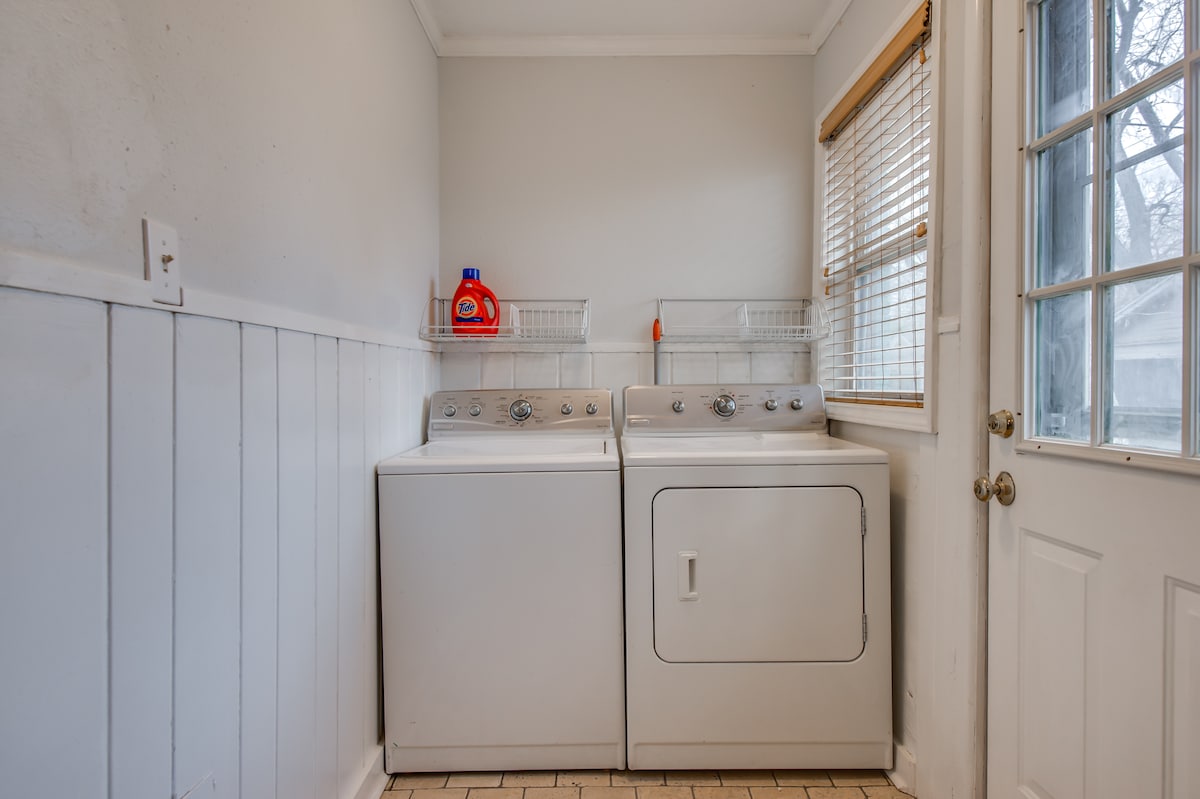 A laundry room is equipped with a washing machine and a dryer, both in white. Shelving above the appliances provides storage for laundry supplies. Natural light enters through a window covered by wooden blinds, and a door with glass panels leads to the outside.