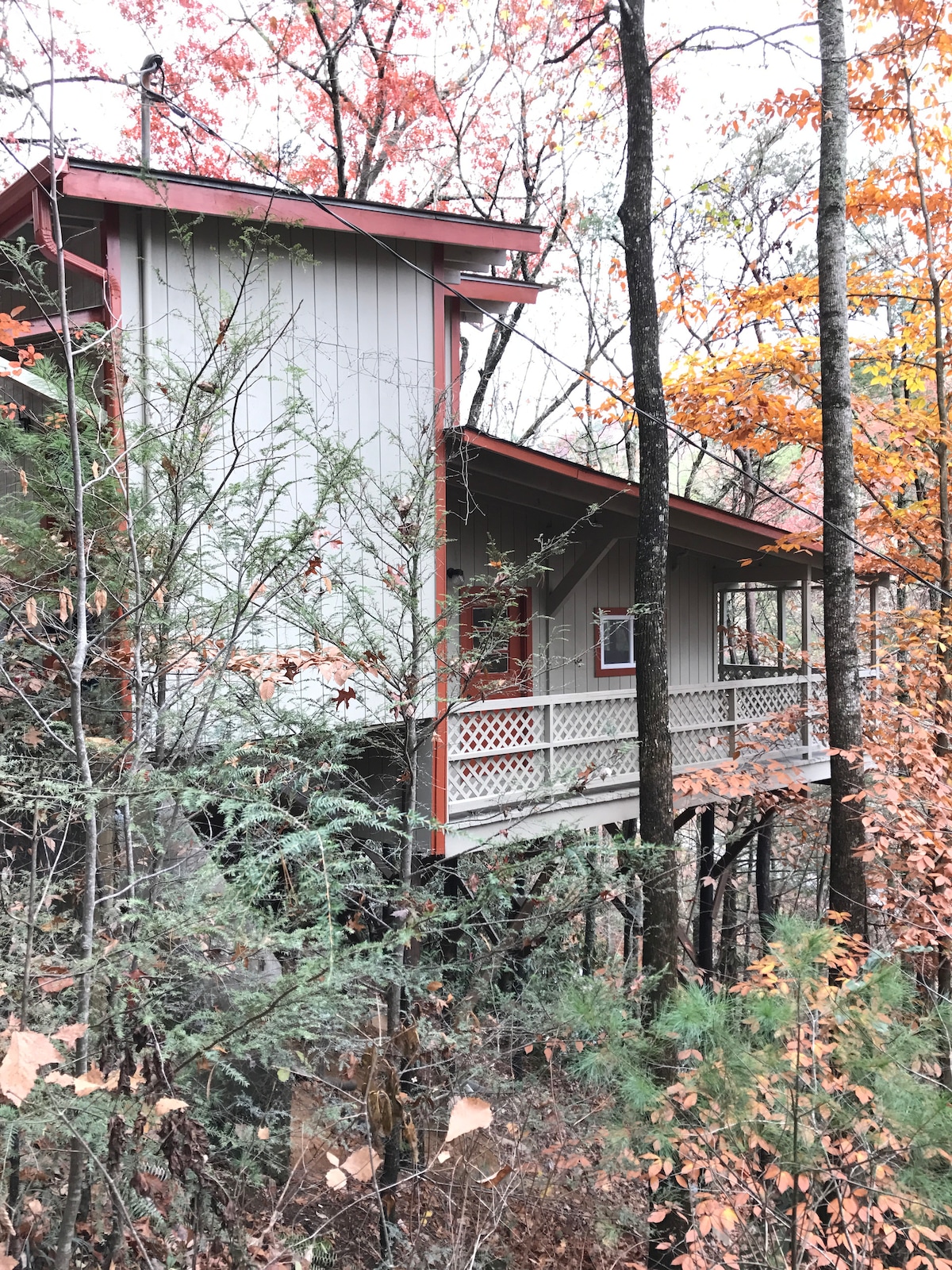 The cabin is elevated among trees, showcasing a split-level design with a red roof and balcony. Autumn foliage in vibrant colors surrounds the structure, enhancing the natural setting. The porch features a railing, inviting relaxation in a serene, wooded environment.