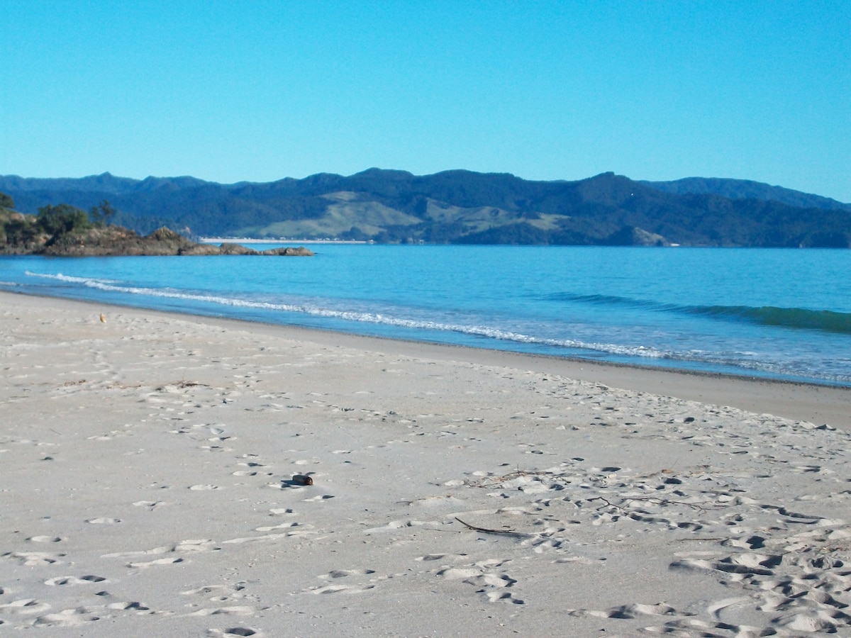 An expansive beach stretches across the foreground, featuring soft sand and gentle waves lapping at the shore. In the distance, rolling hills and vibrant blue waters create a serene coastal landscape under clear skies.