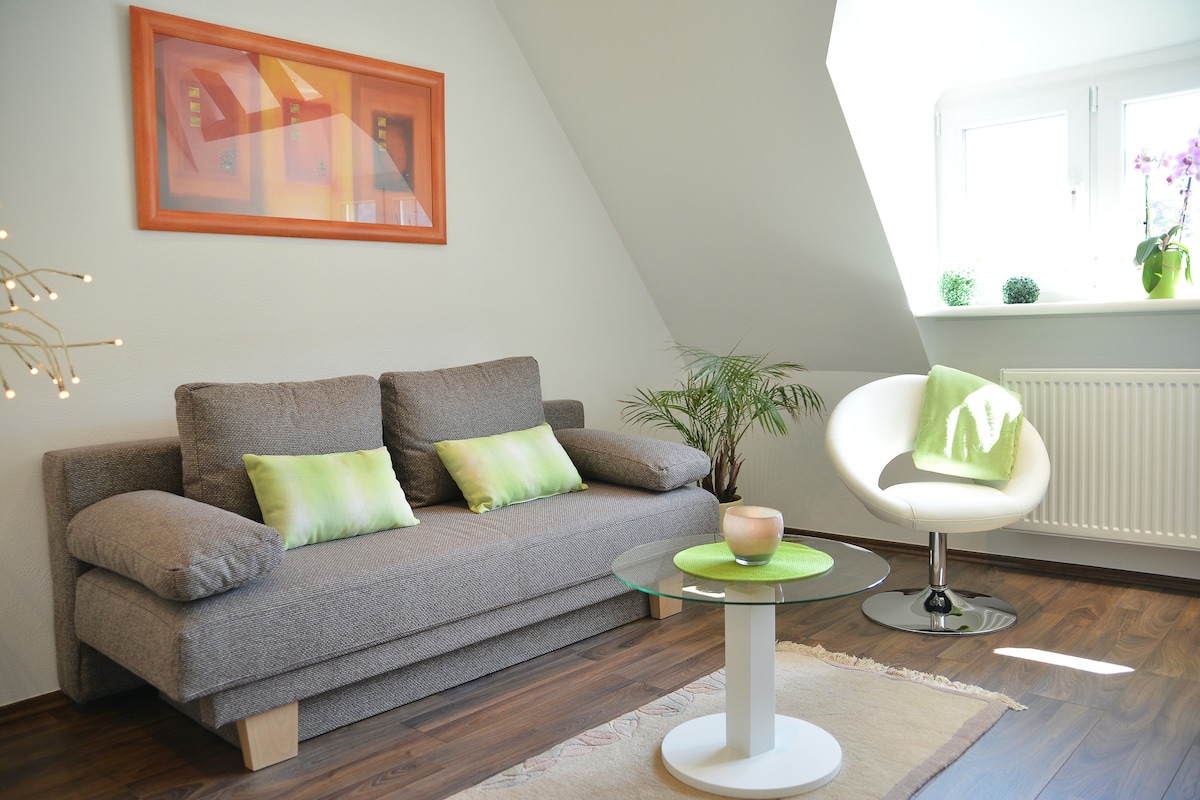 A comfortably furnished living area is shown, featuring a gray sofa with green accent pillows. A round glass table is situated in front, and a white chair with a green throw adds a pop of color. Natural light comes in through two corner windows.